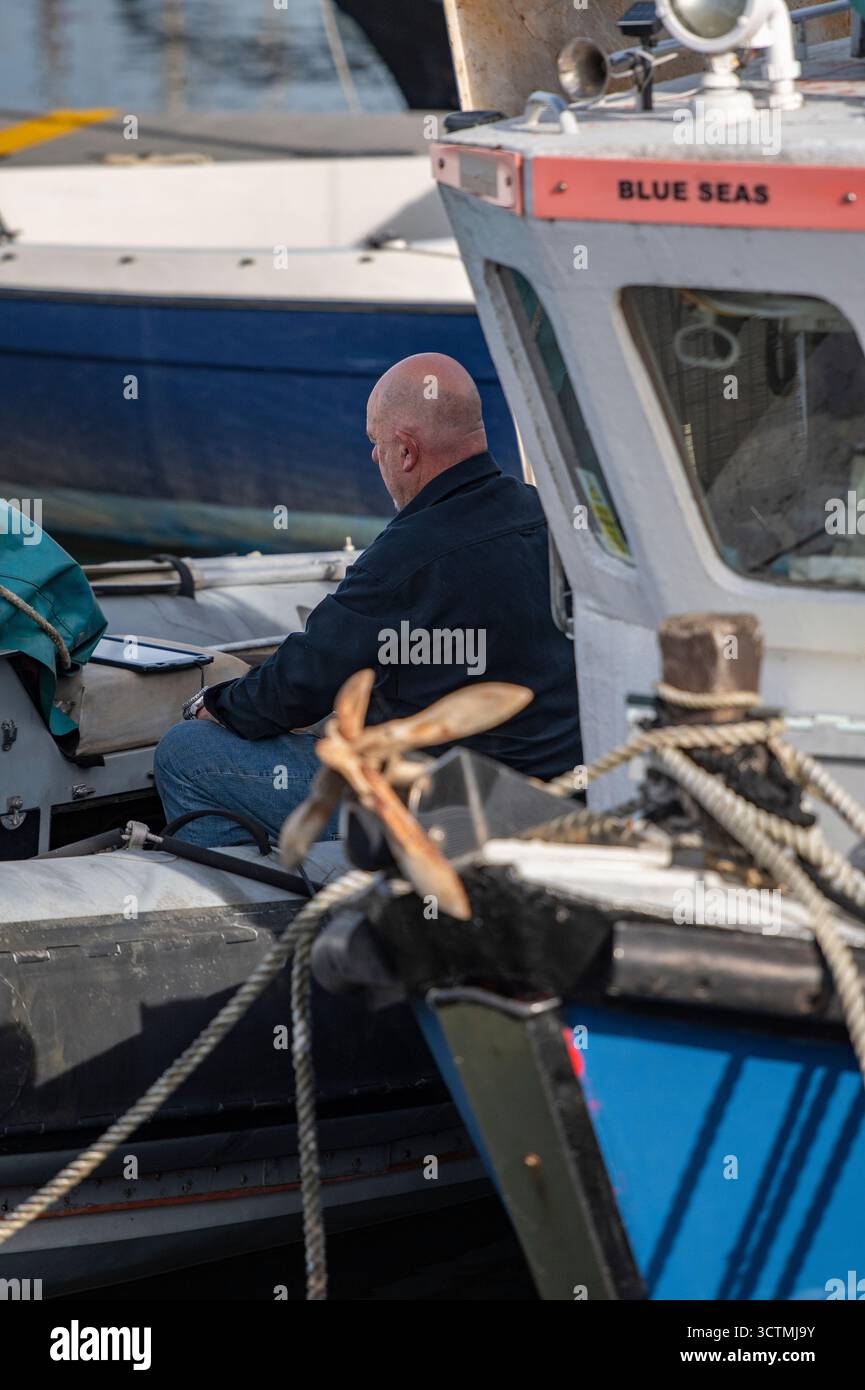 man sitting on a boat working in a marina or harbour. Fisherman sitting on his boat repairing nets and equipment in a port or harbour Stock Photo