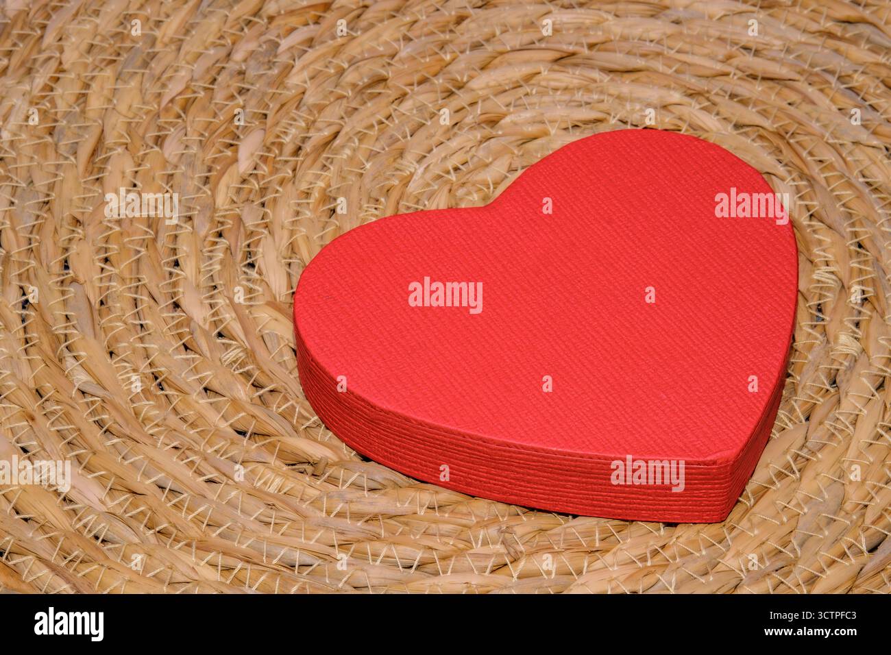Red heart-shaped paper box placed on a woven straw mat. Symbol of love, care and affection with rustic background and simple romantic decoration. Stock Photo