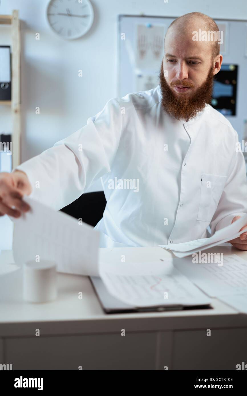 bearded doctor reads paper documents in clinic while working in hospital office. general practitioner studies patient's treatment results. copy space Stock Photo