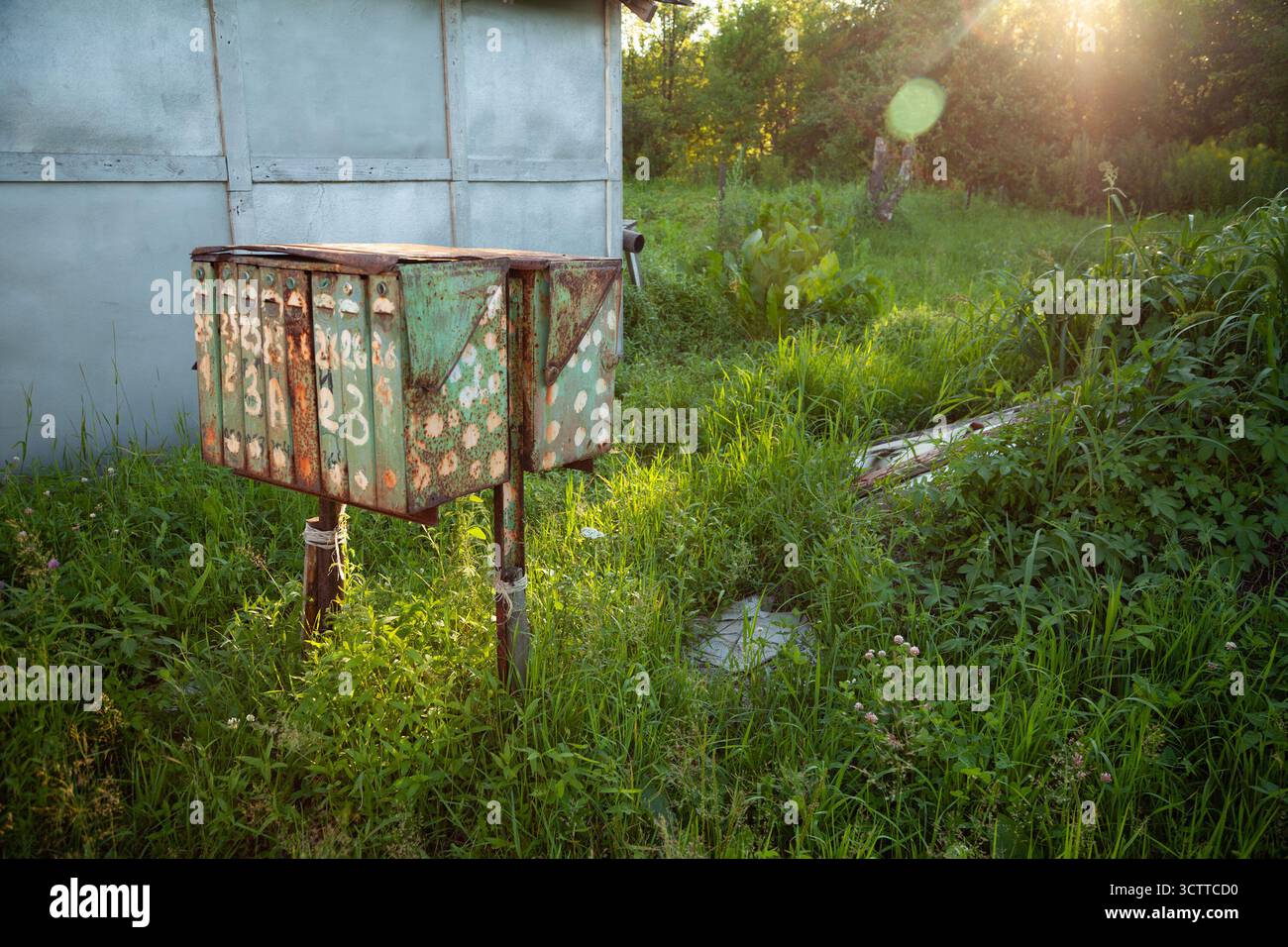 An old, rusty postbox stands weathered in a quiet Russian village, surrounded by green grass. A group of worn metal mailboxes with peeling paint - Stock Image