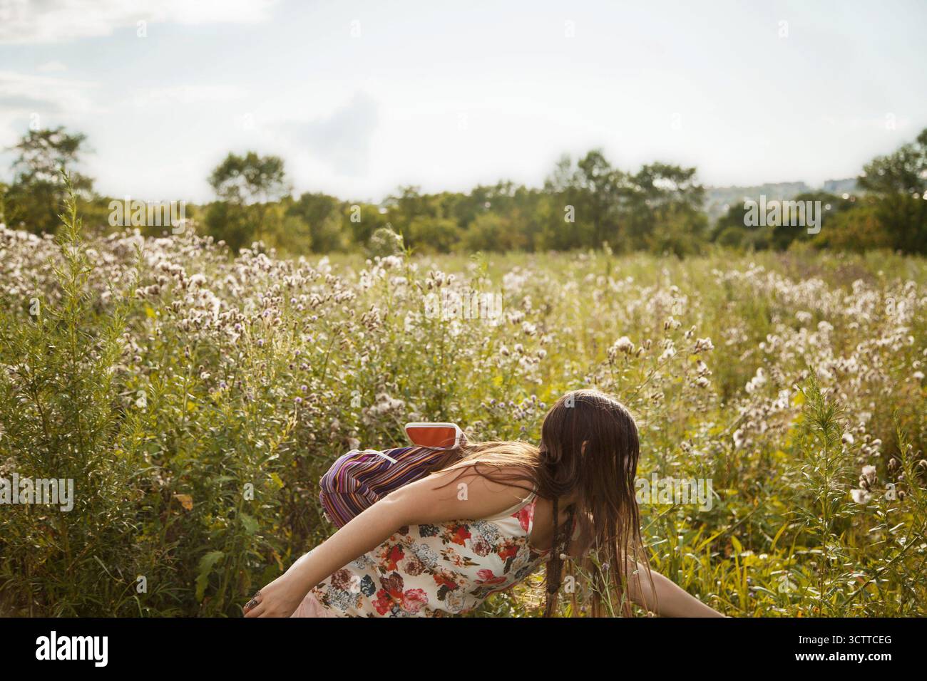 A young woman with long hair bends toward the grass, reaching out to touch a wildflower in the soft autumn light. A European girl with a backpack - Stock Image