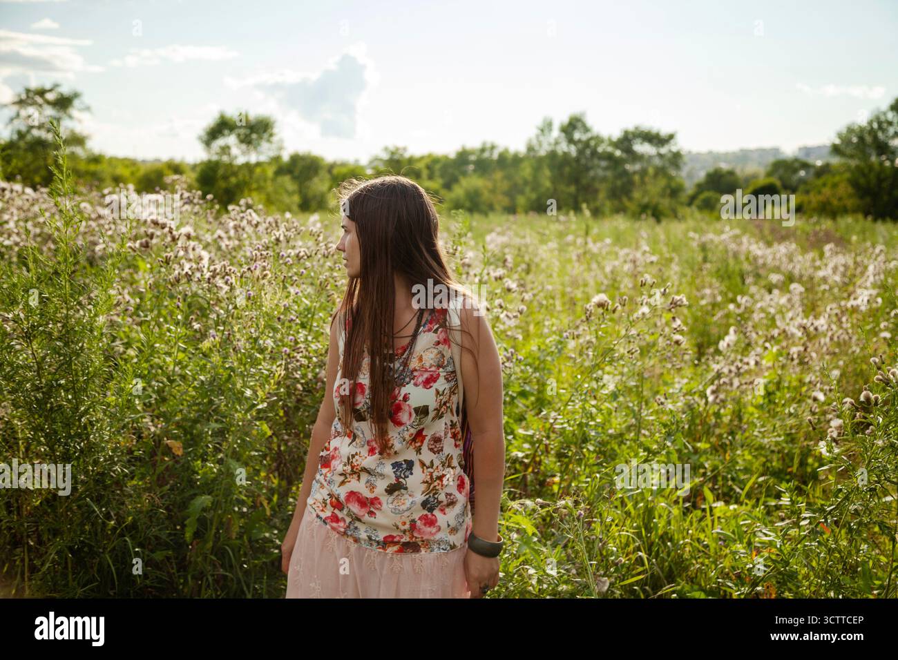 A young woman with long hair stands in a grassy field, looking to the side as wildflowers sway around her. She gazes into the distance, lost in though - Stock Image