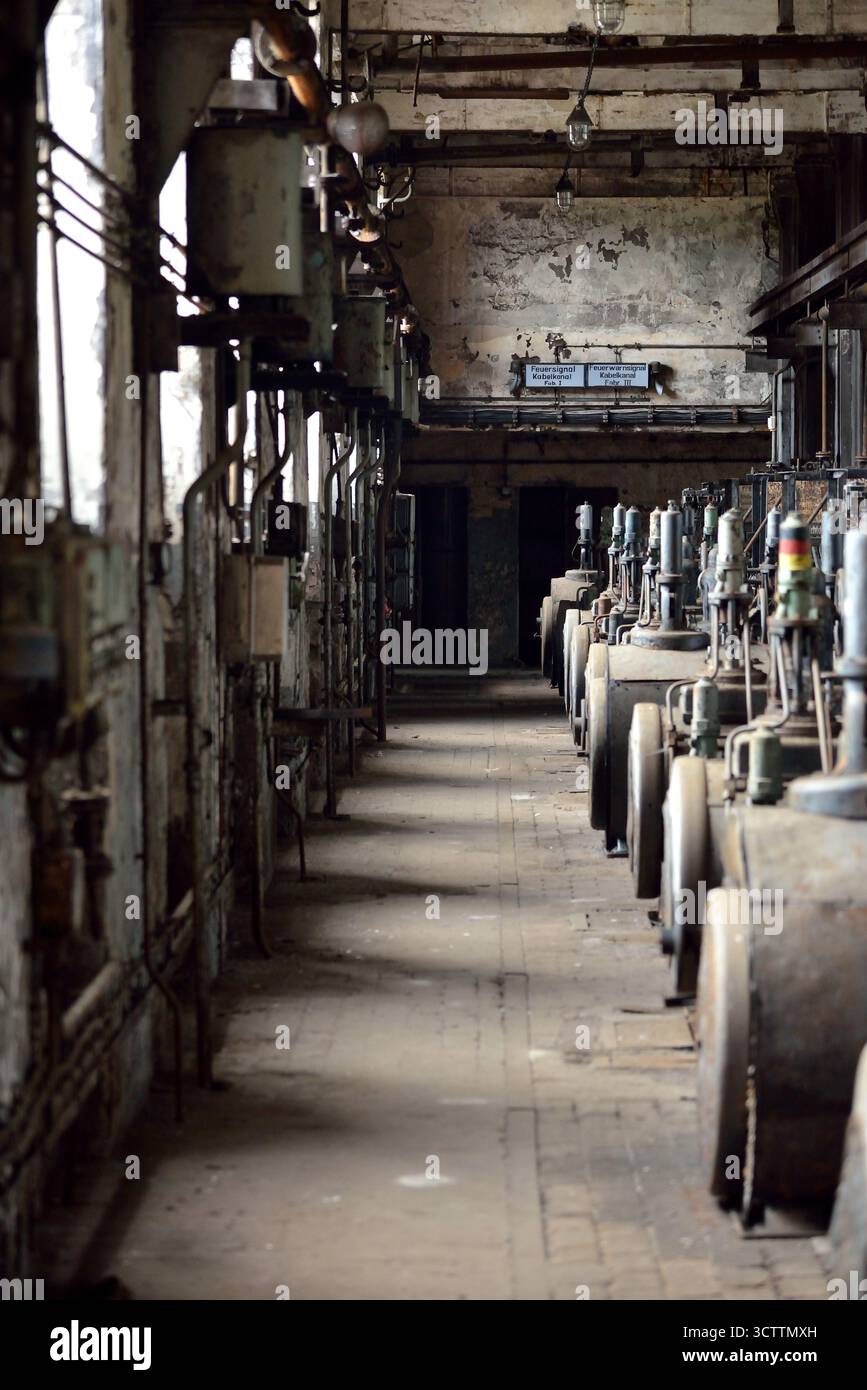 An abandoned corridor in an old, decaying factory hall with a long row of dusty, decommissioned machines Stock Photo