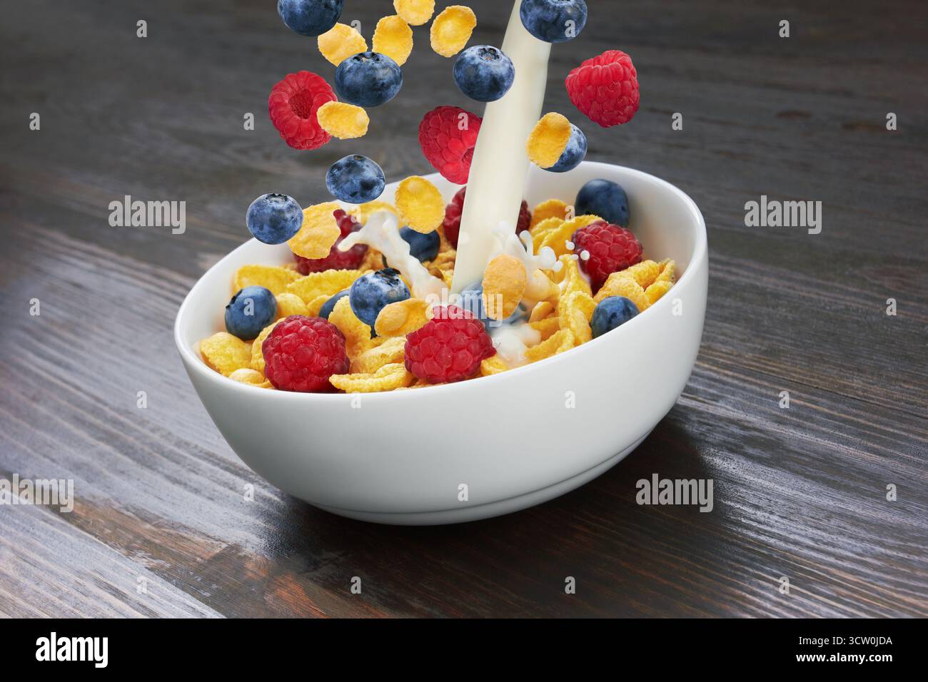 Falling corn flakes with fresh berries and pouring milk on wooden background Stock Photo