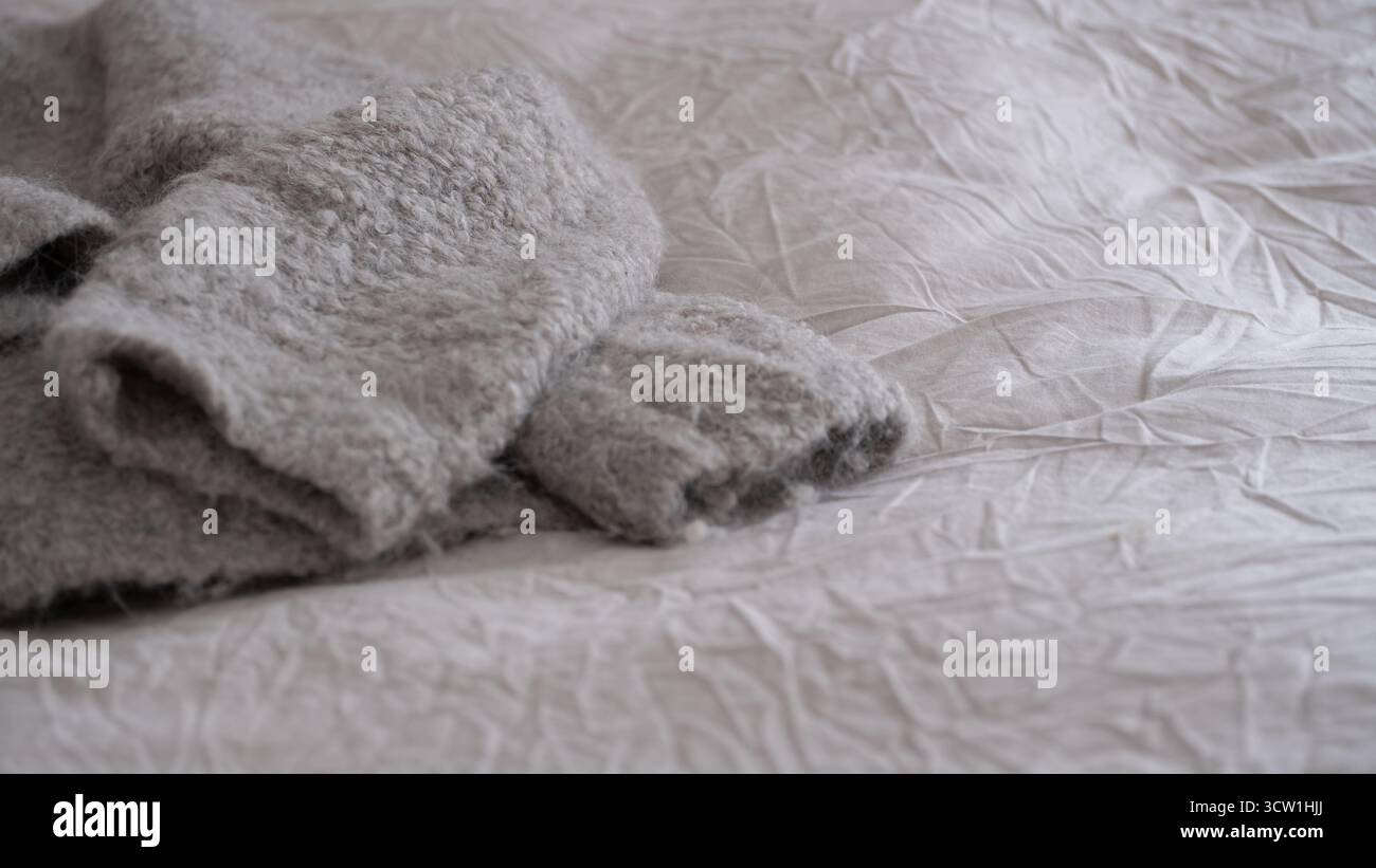 Cozy soft gray wool alpaca sweater lying on a wrinkled light fabric surface. Natural minimal lifestyle composition evoking warmth, comfort, and hygge Stock Photo