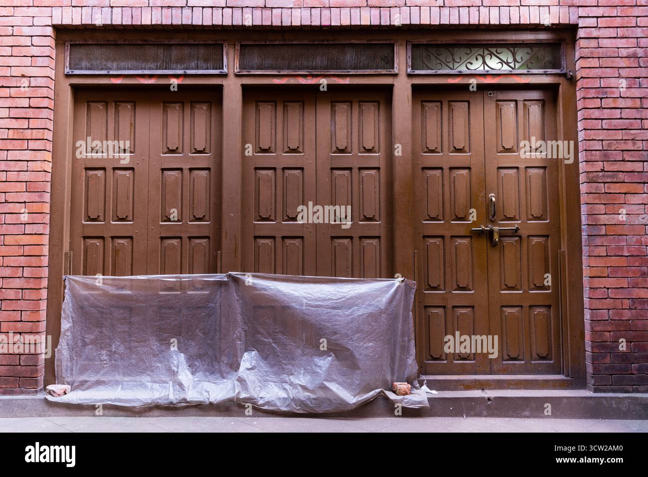 Brick building facade with three large wooden double doors, intricate panels, and weathered textures, partially covered construction materials Stock Photo