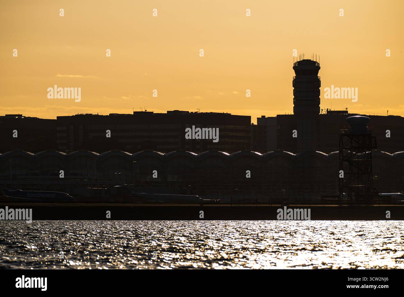 Reagan National Airport Control Tower At Sunset Washington DC // WASHINGTON DC — The control tower and terminal of Ronald Reagan Washington National Airport (DCA) are silhouetted against the setting sun. This major airport, serving the Washington metropolitan area, is located on the Potomac River. The image is captured from Hains Point, a peninsula within East Potomac Park, offering views of the airport, which is a hub for American Airlines and Southwest Airlines. Stock Photo