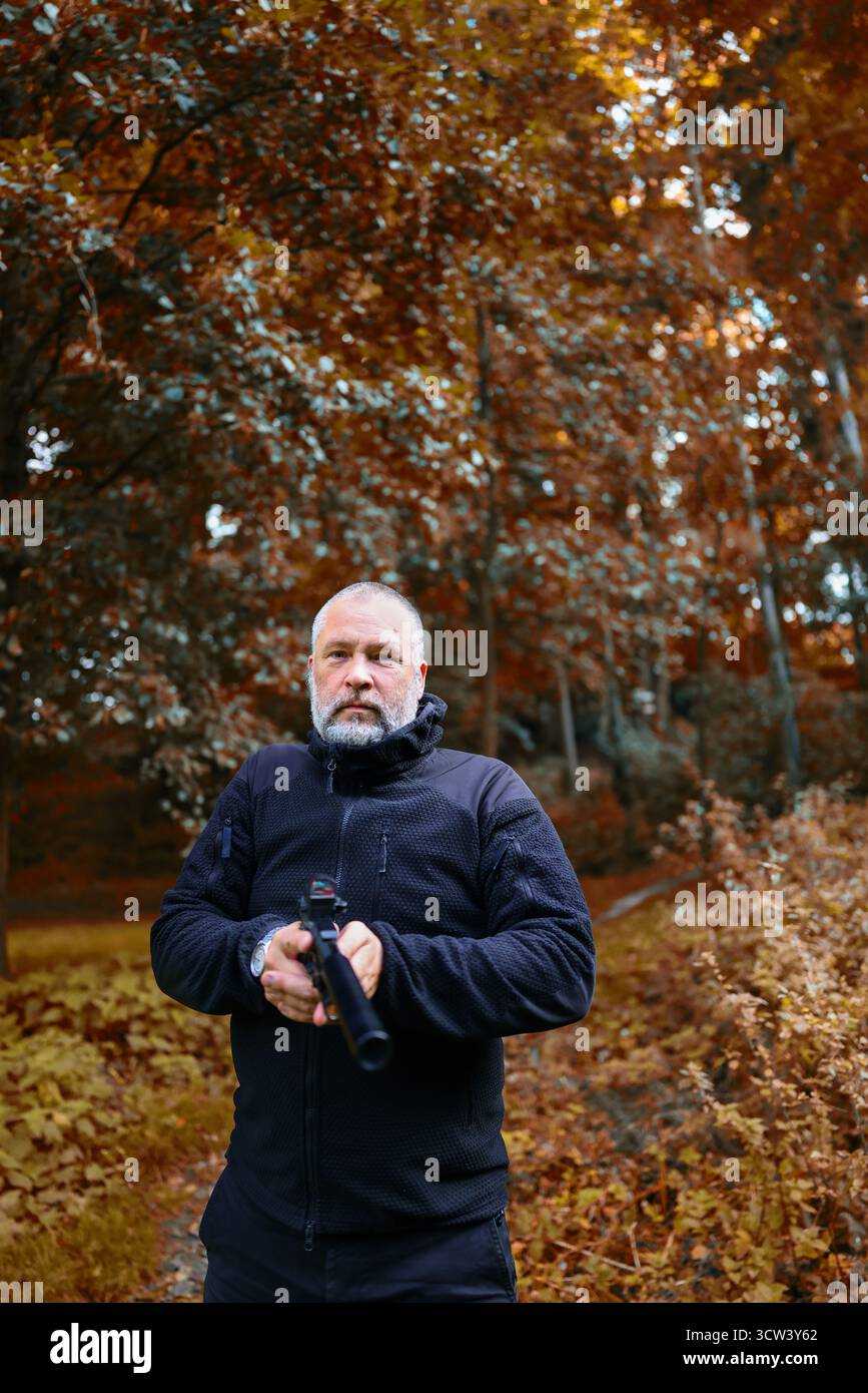 Bearded man in black jacket with gun, security concept in nature setting. copy space Stock Photo
