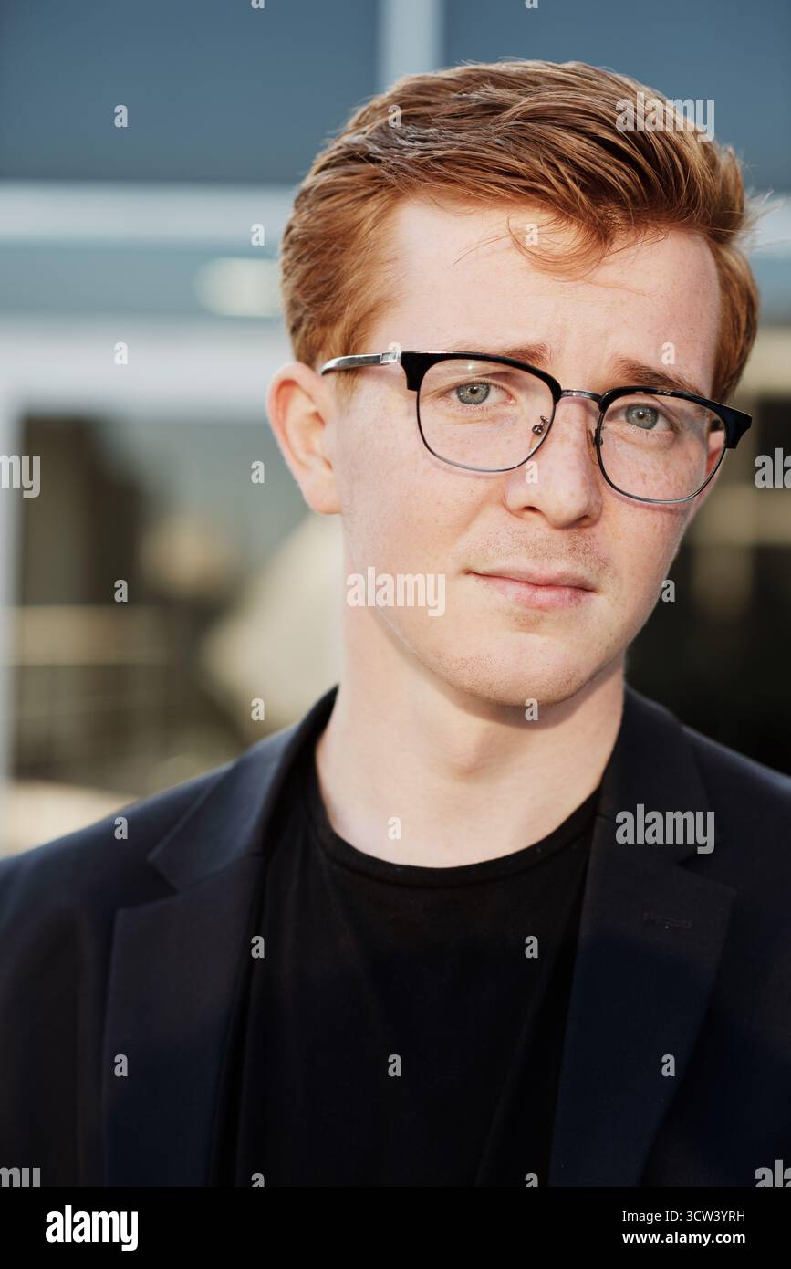 Chest-up portrait of young confident ginger Caucasian man wearing glasses and black clothes looking at camera with serious face expression Stock Photo