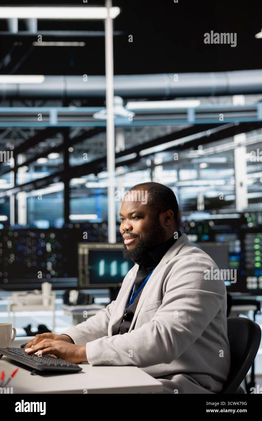 Upbeat data center IT expert uses PC to update servers tech, configuring gear using software. Jolly server room programmer reviewing hardware analytics, ensuring reliable network operations Stock Photo