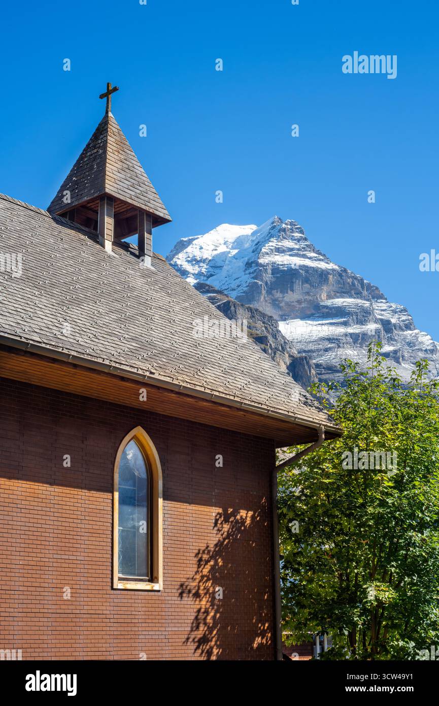 A church with a wooden roof and steeple against a snowy mountain backdrop in alpine village of Murren, Switzerland. Stock Photo