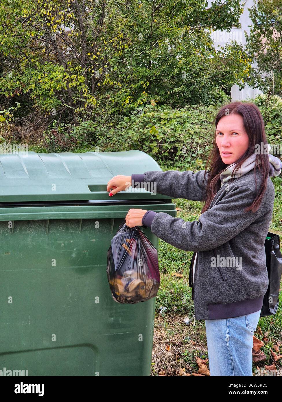 A woman throws a bag of trash into a trash bin. Outdoor waste disposal. - Smartphone Captured Stock Image