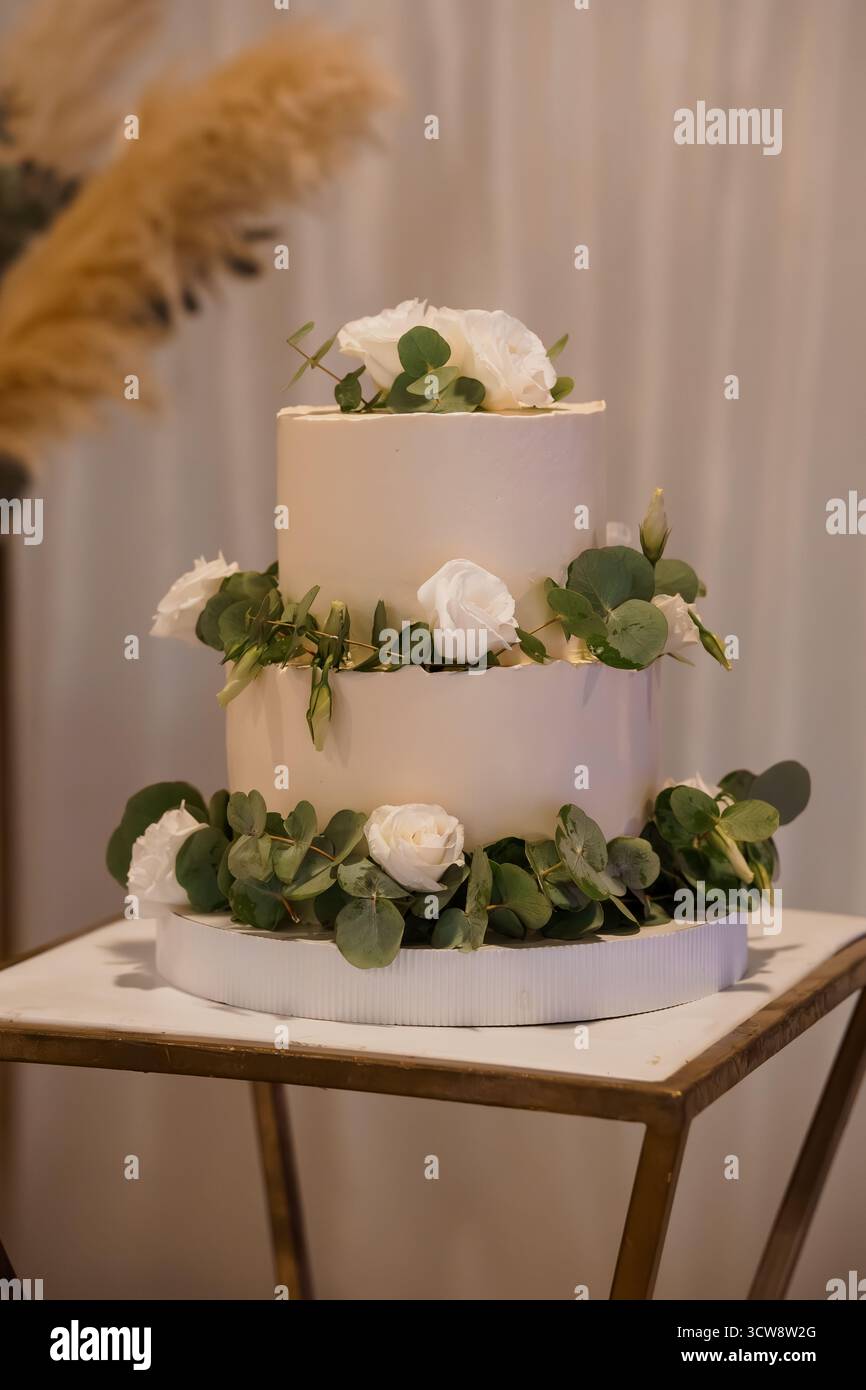 A beautifully decorated two-tier wedding cake stands on a wooden stand. adorned with white roses and eucalyptus greenery, this dessert symbolizes love Stock Photo