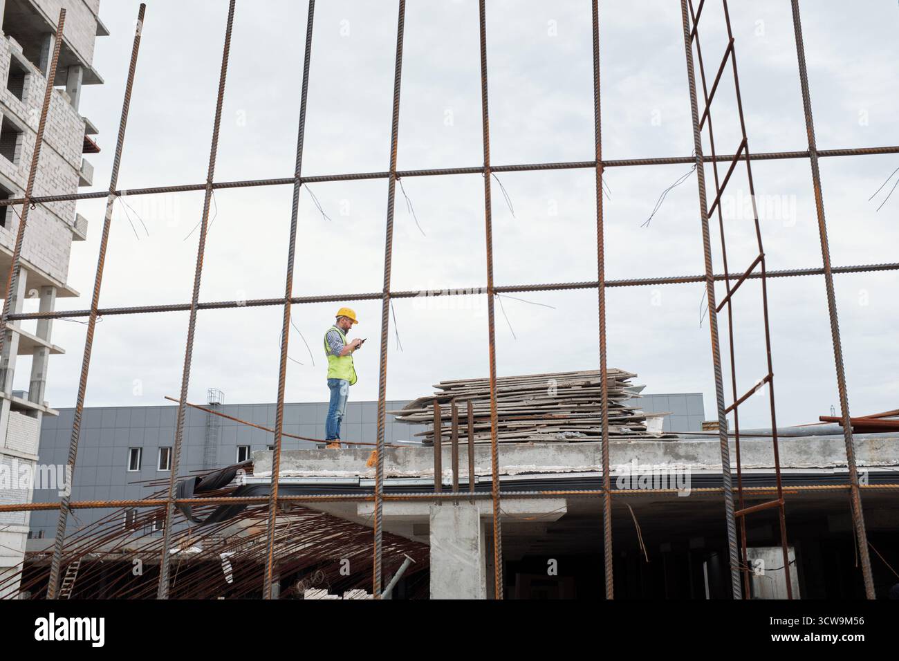 Caucasian middle aged man wearing safety helmet standing on construction site using smartphone, surrounded by steel framework and building materials during workday Stock Photo