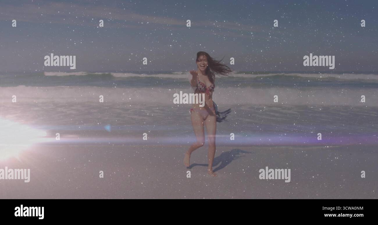 Running adult woman wearing floral bikini holding towel on sandy beach, sun flare and ocean waves Stock Photo