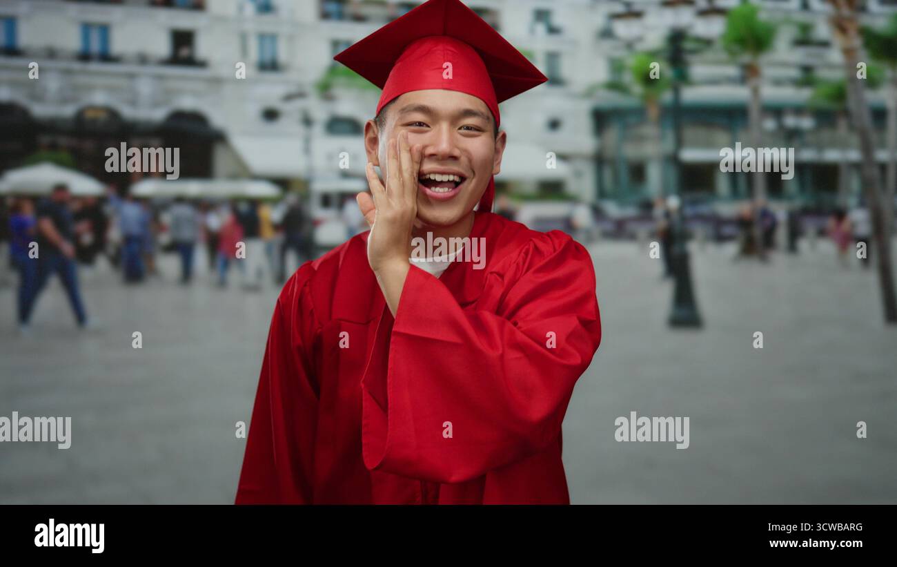 Secretively Young man in red graduation gown smiles and gestures secretively on a ...