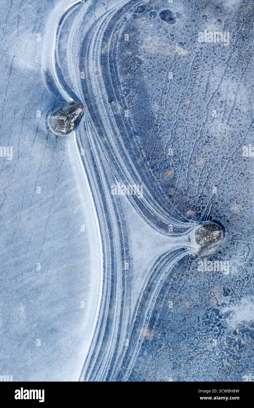 Two small stones break through a sea of swirling frozen ice, creating an abstract pattern of curves and arcs - Stock Image