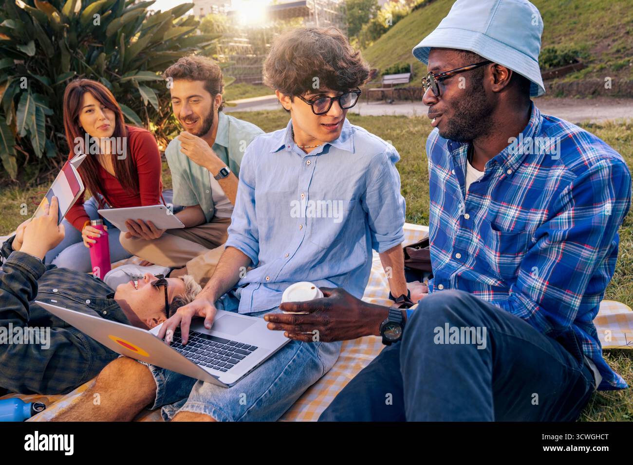 A group of happy young multi-ethnic students collaborates on a project, using a laptop and tablets while studying together on a blanket in a park duri Stock Photo