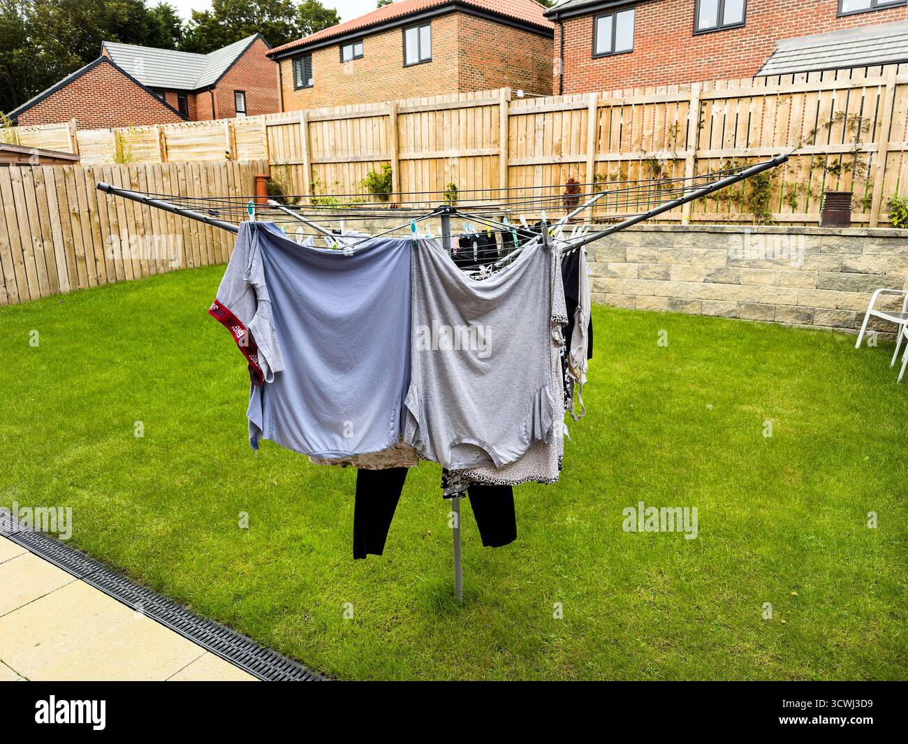 A rotary clothesline in a backyard with various clothes hung to dry, surrounded by fenced grassy area and residential homes. uk - Smartphone Captured Stock Image