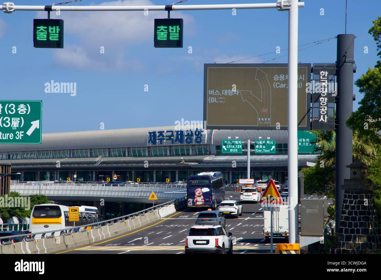 Traffic flows toward Jeju International Airport’s terminal on elevated ramps and multilane roads, with overhead signs sending buses and cars to depart Stock Photo