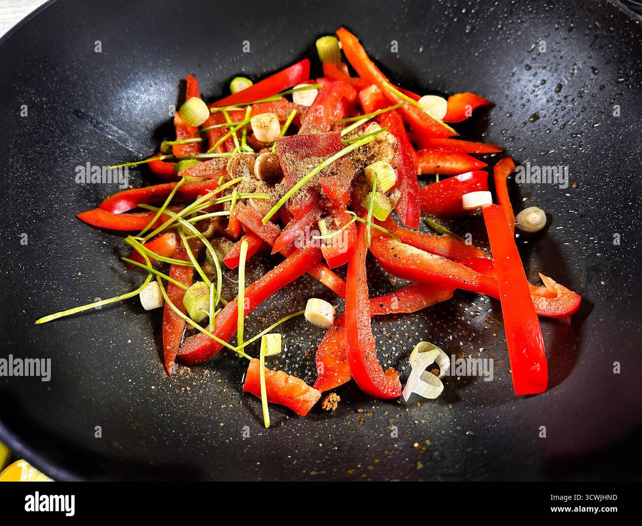 Slices of fresh red bell pepper and green spring onion being stir-fried in a black frying pan wok with spices and herbs for a flavorful dish - Smartphone Captured Stock Image