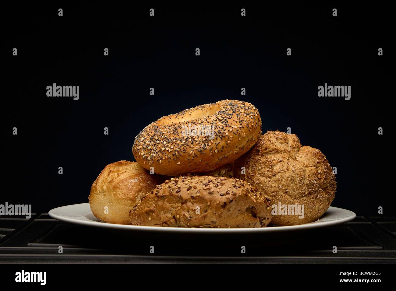 Assorted fresh bread rolls stacked on a white plate against dark background illuminated by purple and cyan light. Modern bakery concept image with art Stock Photo