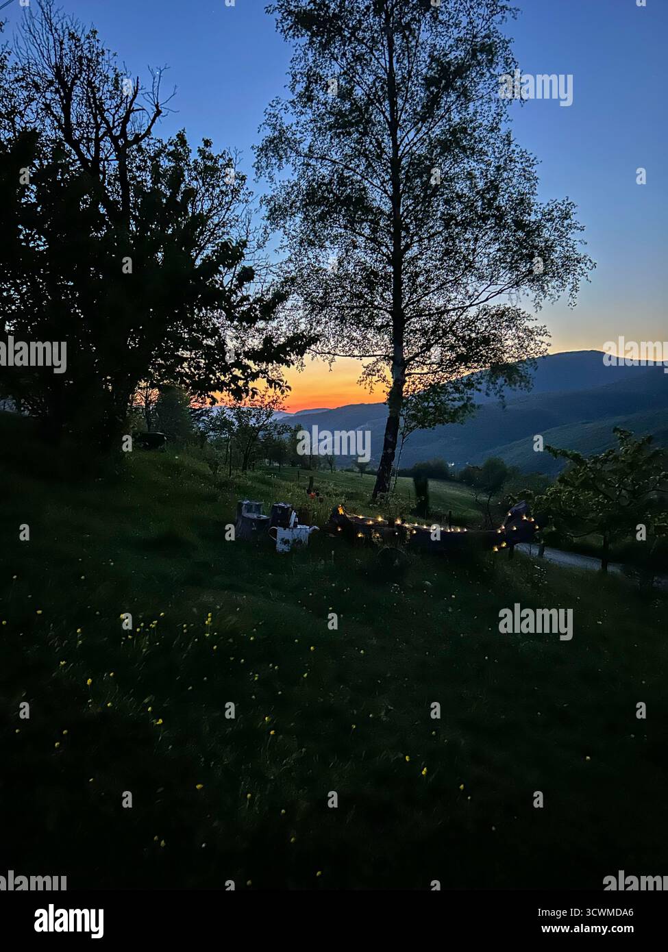 Tranquil mountain landscape at dusk with glowing sunset, trees in silhouette, and soft lights illuminating a rustic countryside setting. - Smartphone Captured Stock Image