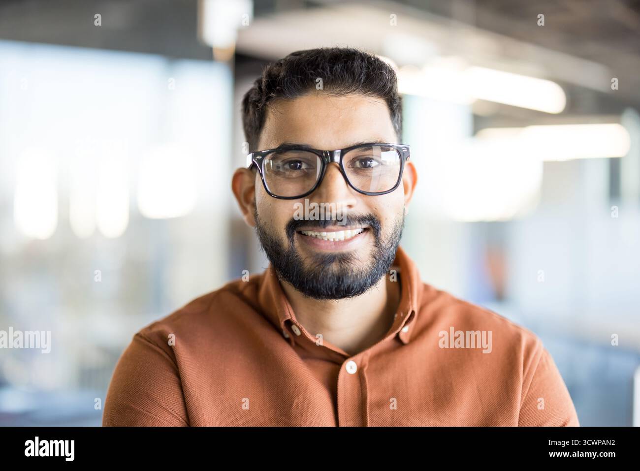 Young indian man with a beard and glasses smiling confidently at the camera, portraying a successful and approachable professional in a modern office environment Stock Photo