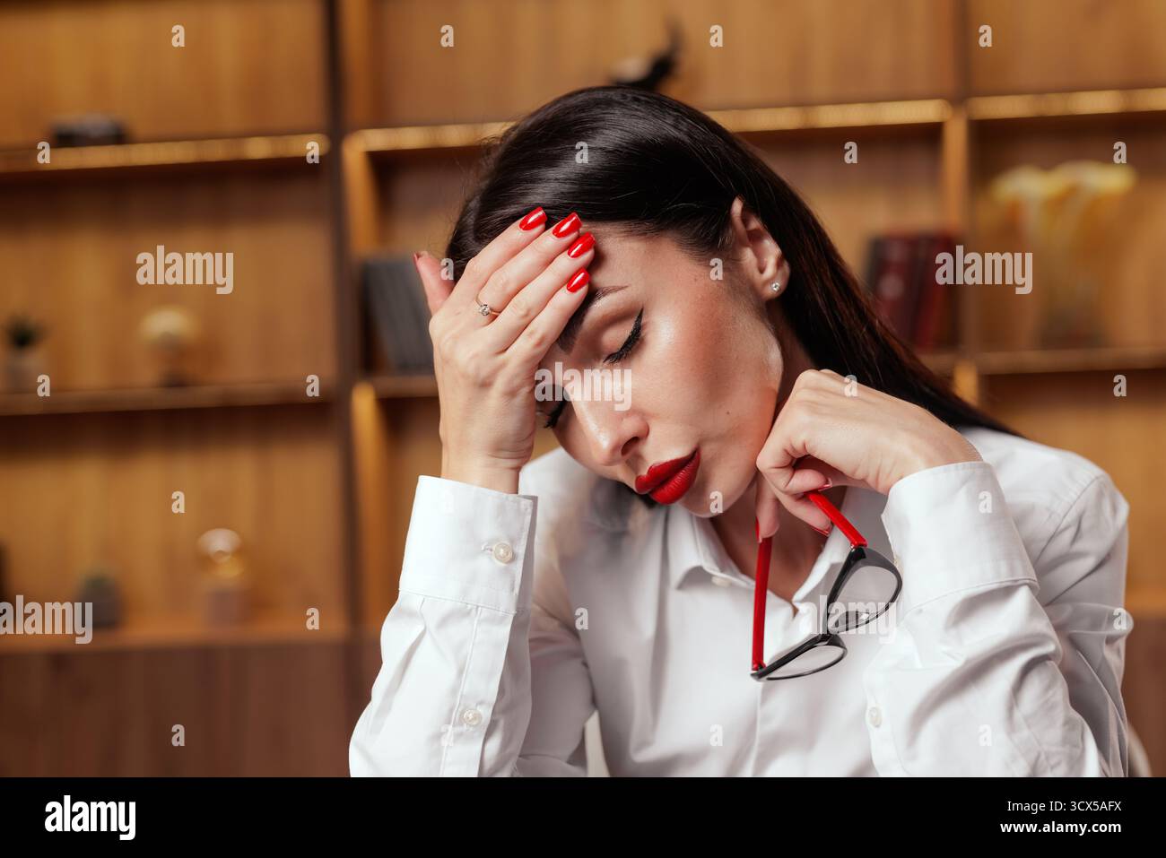 Close view of tired woman holding her head with glasses in hand, illustrating migraine headache and stress linked to triggers and work pressure Stock Photo