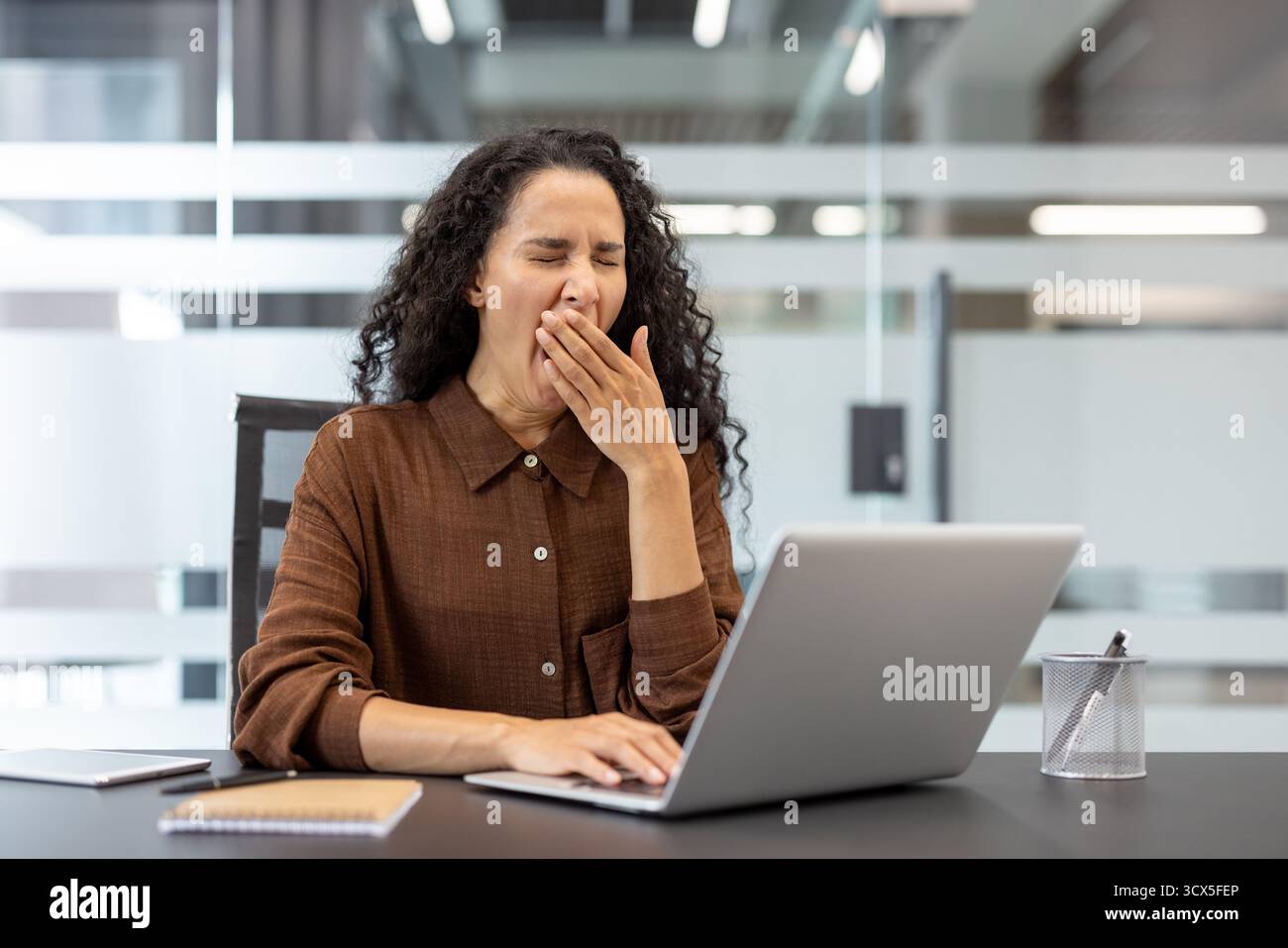 Businesswoman yawning and covering her mouth at a modern office desk, tired and overworked while working on a laptop, showing burnout, fatigue and low energy during long hours Stock Photo