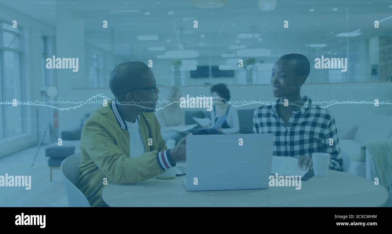 Two colleagues discussing project while gesturing toward laptop in open-plan office, with paperwork Stock Photo