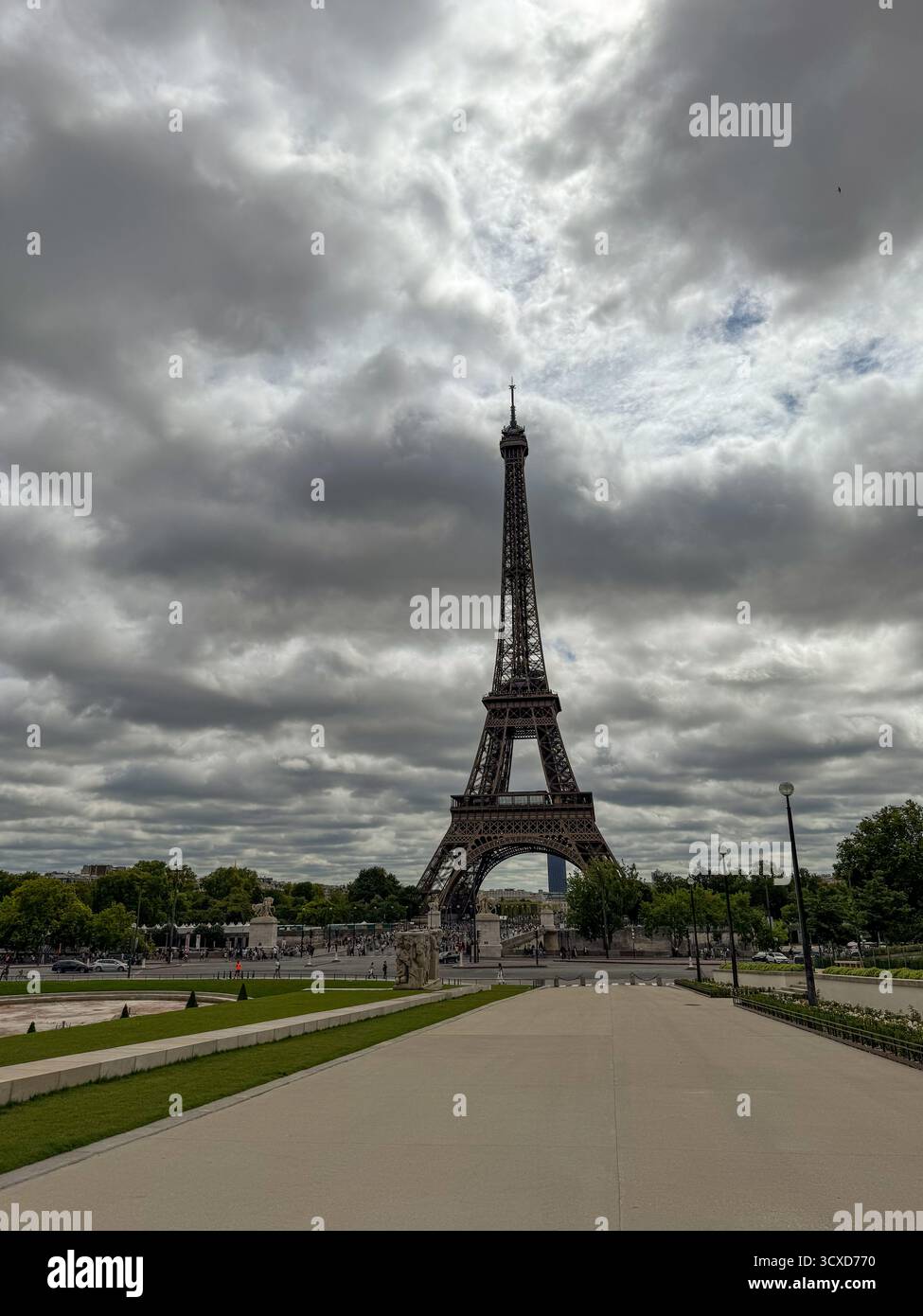 a low angle vertical view of the Eiffel Tower in Paris, France, rising above a wide empty pathway and green lawns under a dramatic cloudy sky. - Smartphone Captured Stock Image