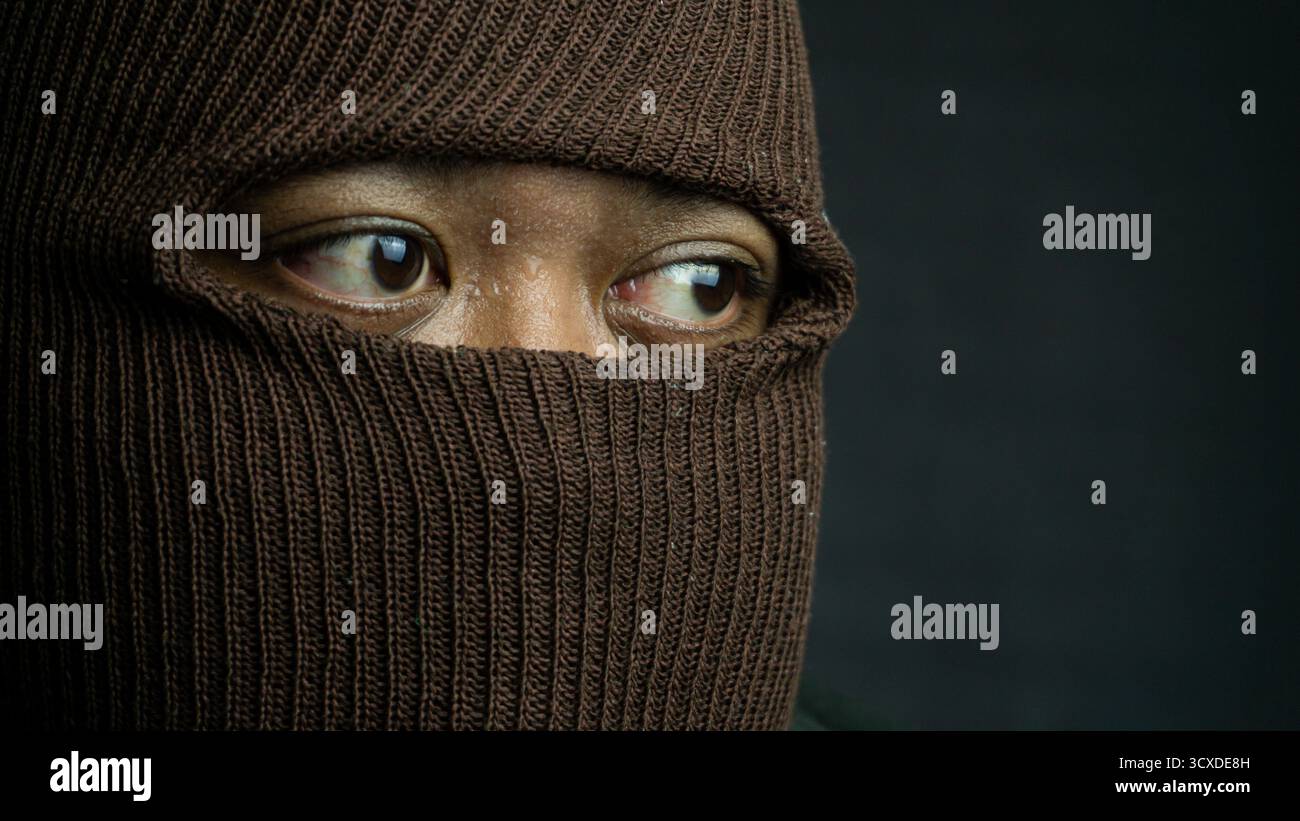 Close-up portrait of man with brown cloth mask covering face except eyes, intense and secretive look with sharp shadows on black background Stock Photo