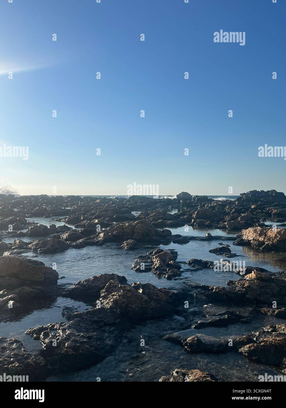 A tranquil view of a rocky shoreline under a clear blue sky. The sunlight reflects off the wet rocks, creating a natural coastal scene. - Smartphone Captured Stock Image