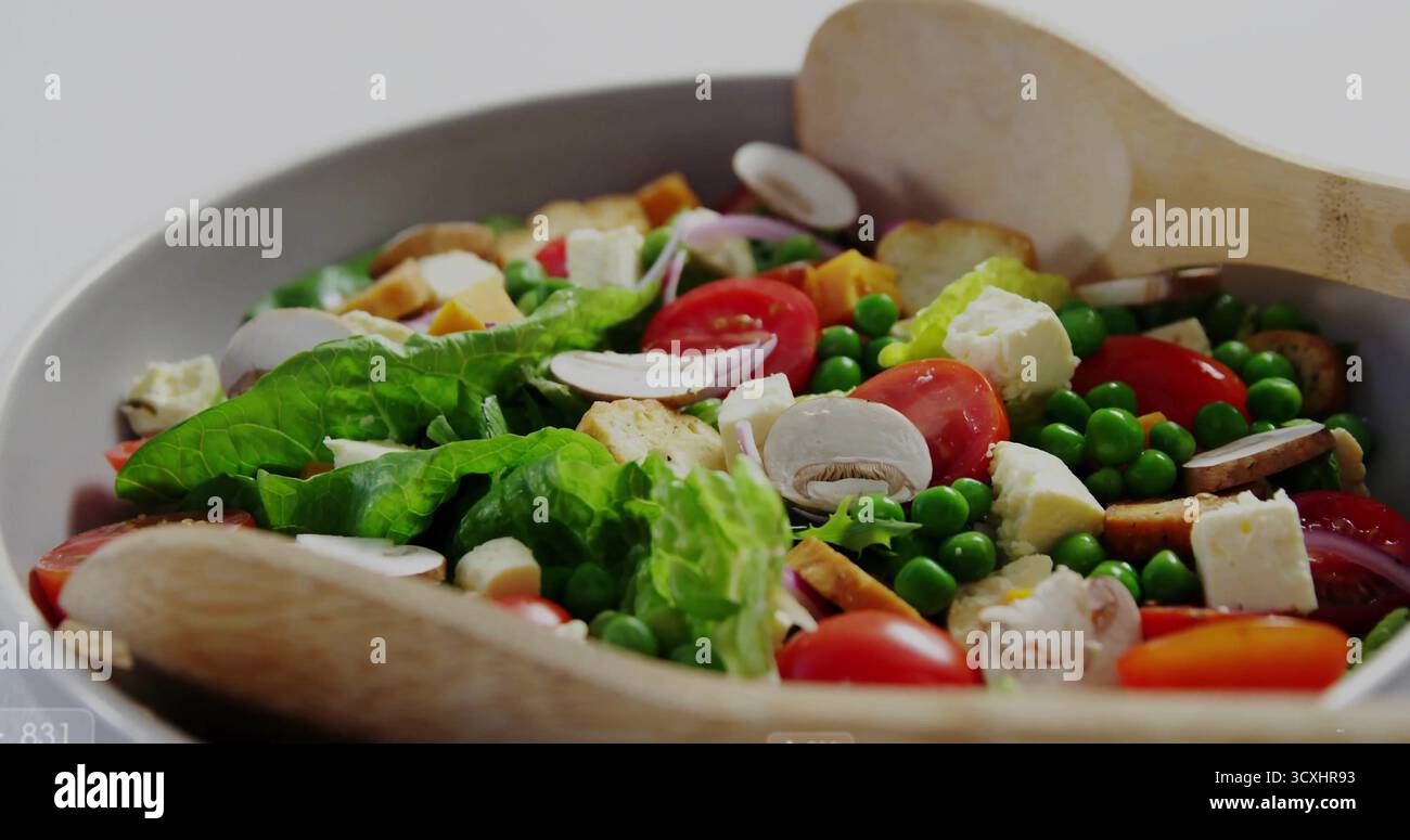 White ceramic bowl resting on light tabletop, with salad, tomatoes, croutons, wooden tongs Stock Photo