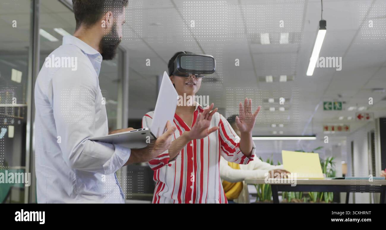 Gesturing woman wearing VR headset in open-plan office with laptops, paper and bar-height table Stock Photo