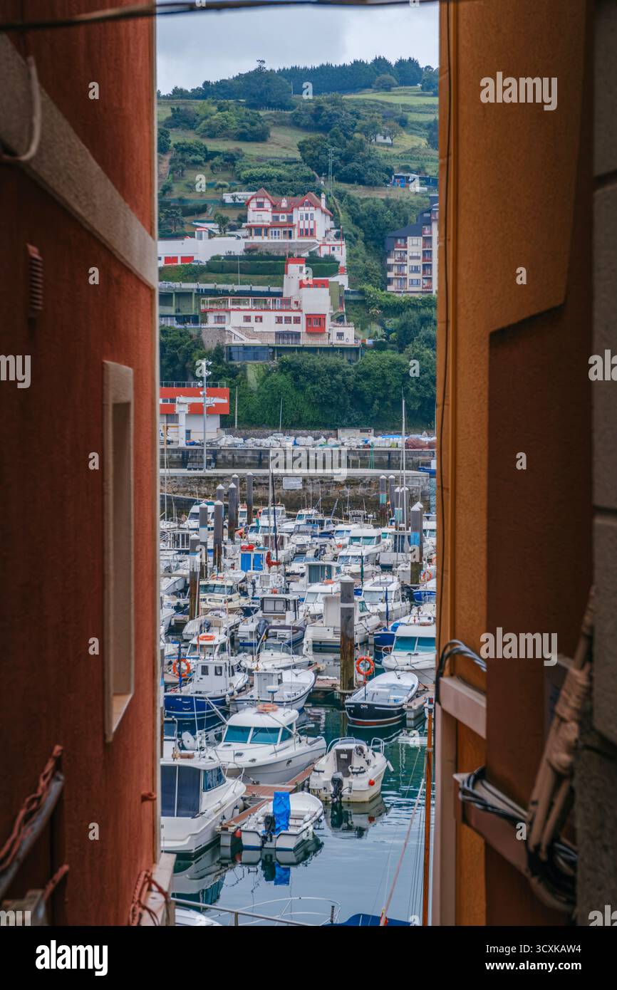 Scenic harbor view framed by Narrow Alley showing fishing boats and traditional white houses on hillside in Bermeo fishing port, Basque Country, Spain Stock Photo