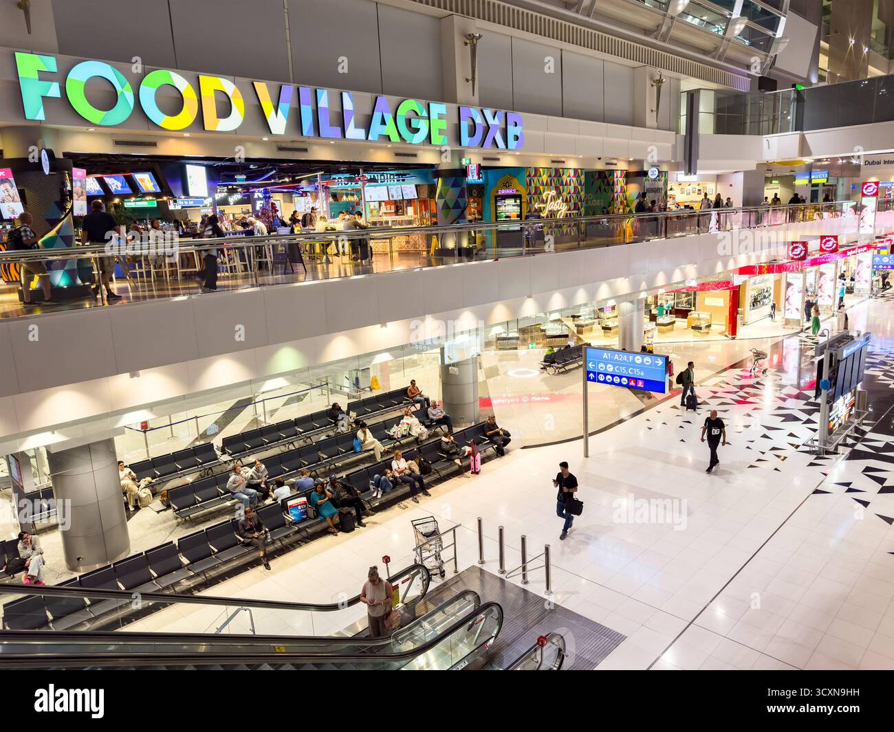 A busy terminal area at Dubai International Airport featuring Food Village DXB and seated travelers on an escalator - Smartphone Captured Stock Image
