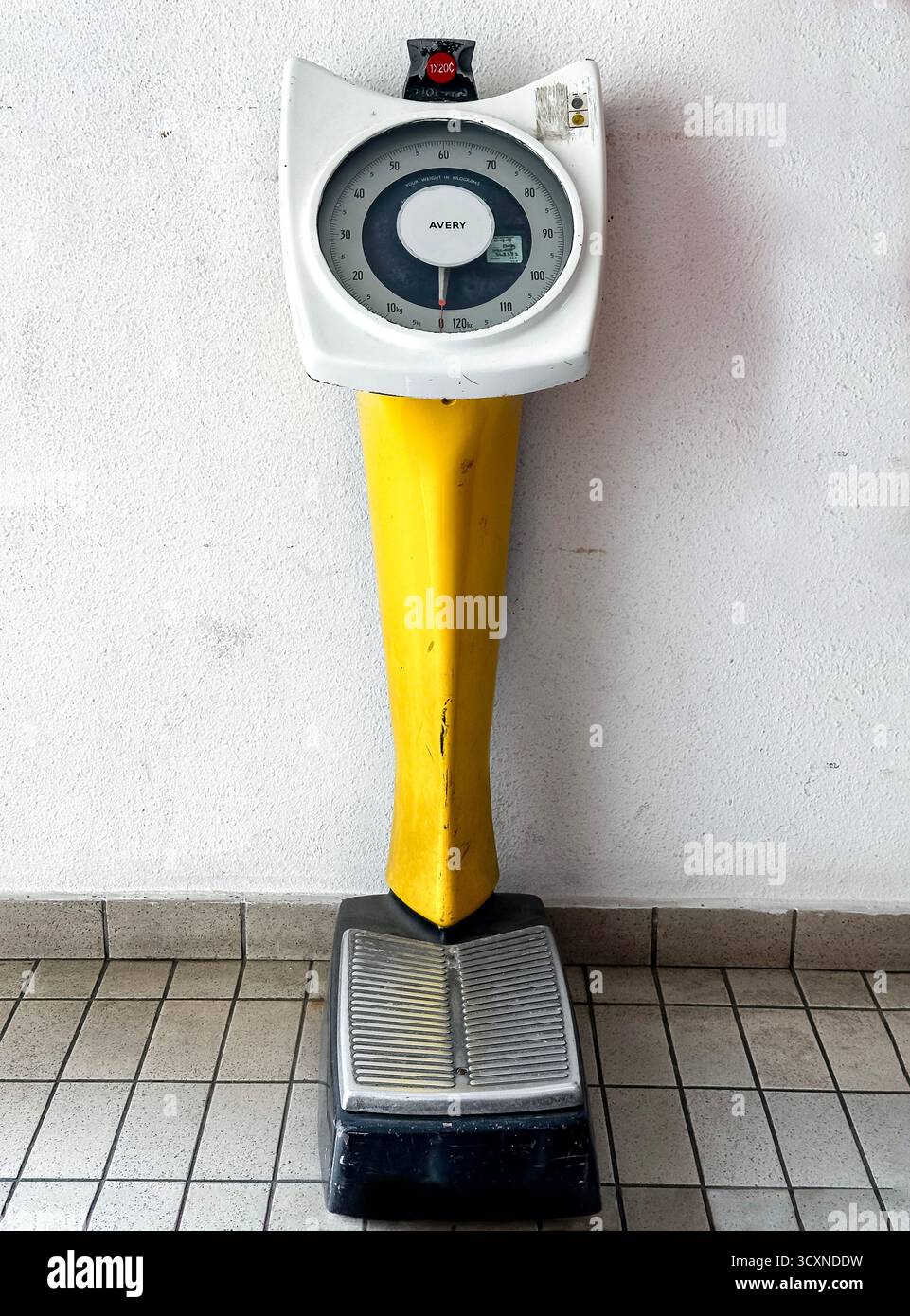 Vintage Avery coin operated weighing scales with a yellow base against a white wall, standing on a tiled floor in public toilet kuala lumpur malaysia - Smartphone Captured Stock Image