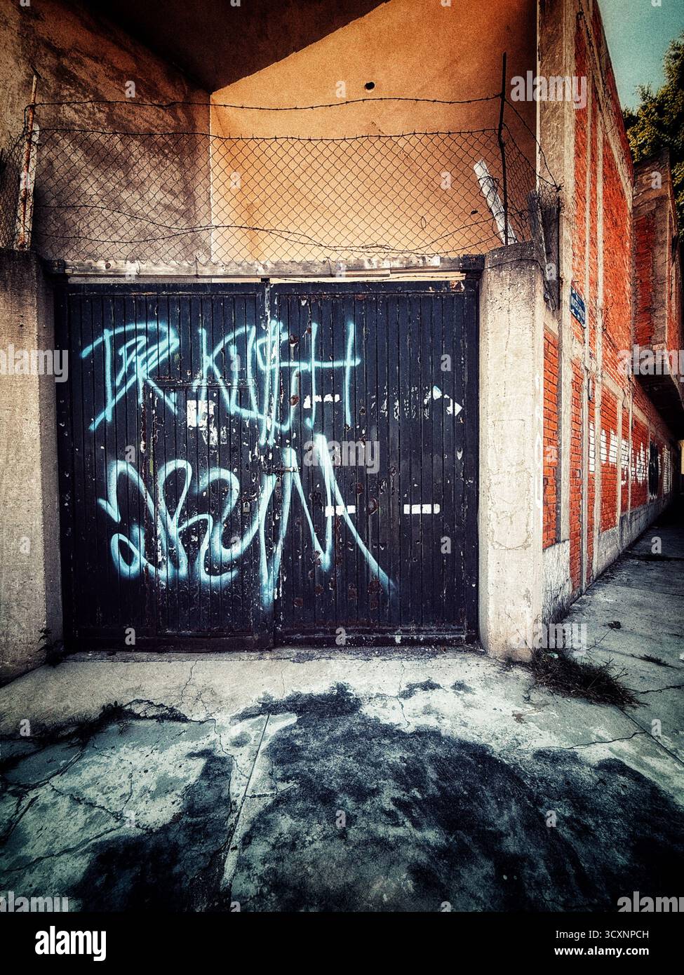 Old garage door with blue graffiti in an urban alley surrounded by brick walls and harsh shadows - Smartphone Captured Stock Image
