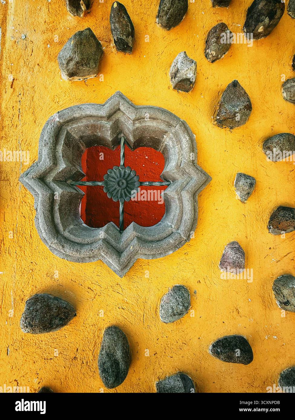 Ornate window with intricate design set in a vibrant yellow wall surrounded by scattered dark stones - Smartphone Captured Stock Image