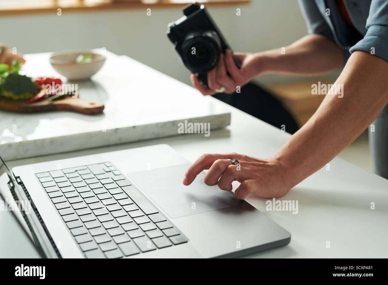 Man Using Laptop While Holding Camera in Kitchen Setting Stock Photo