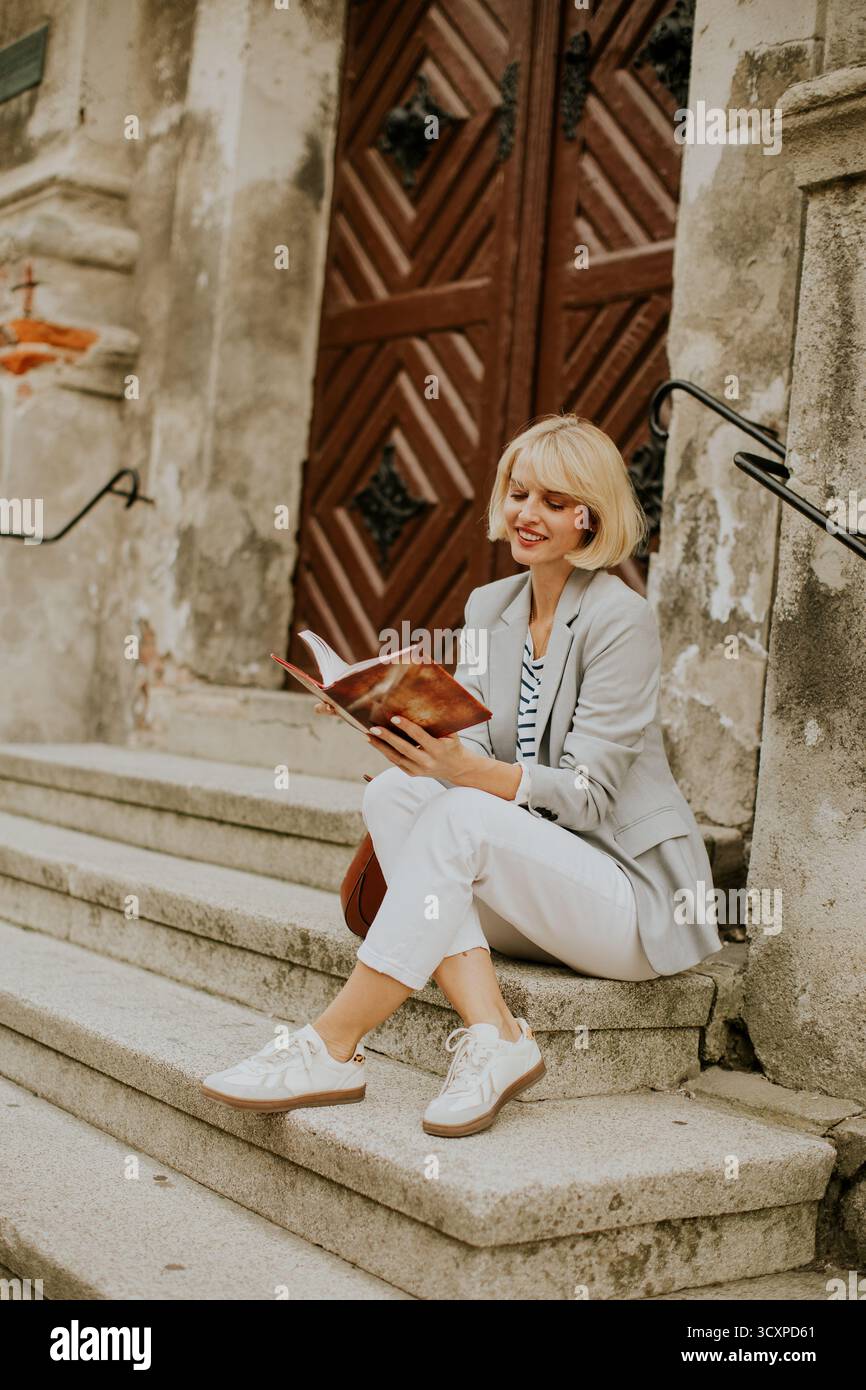 A young woman with short blonde hair sits on the steps of an old building. She reads a book while dressed in a blazer and casual shoes, enjoying a sun Stock Photo