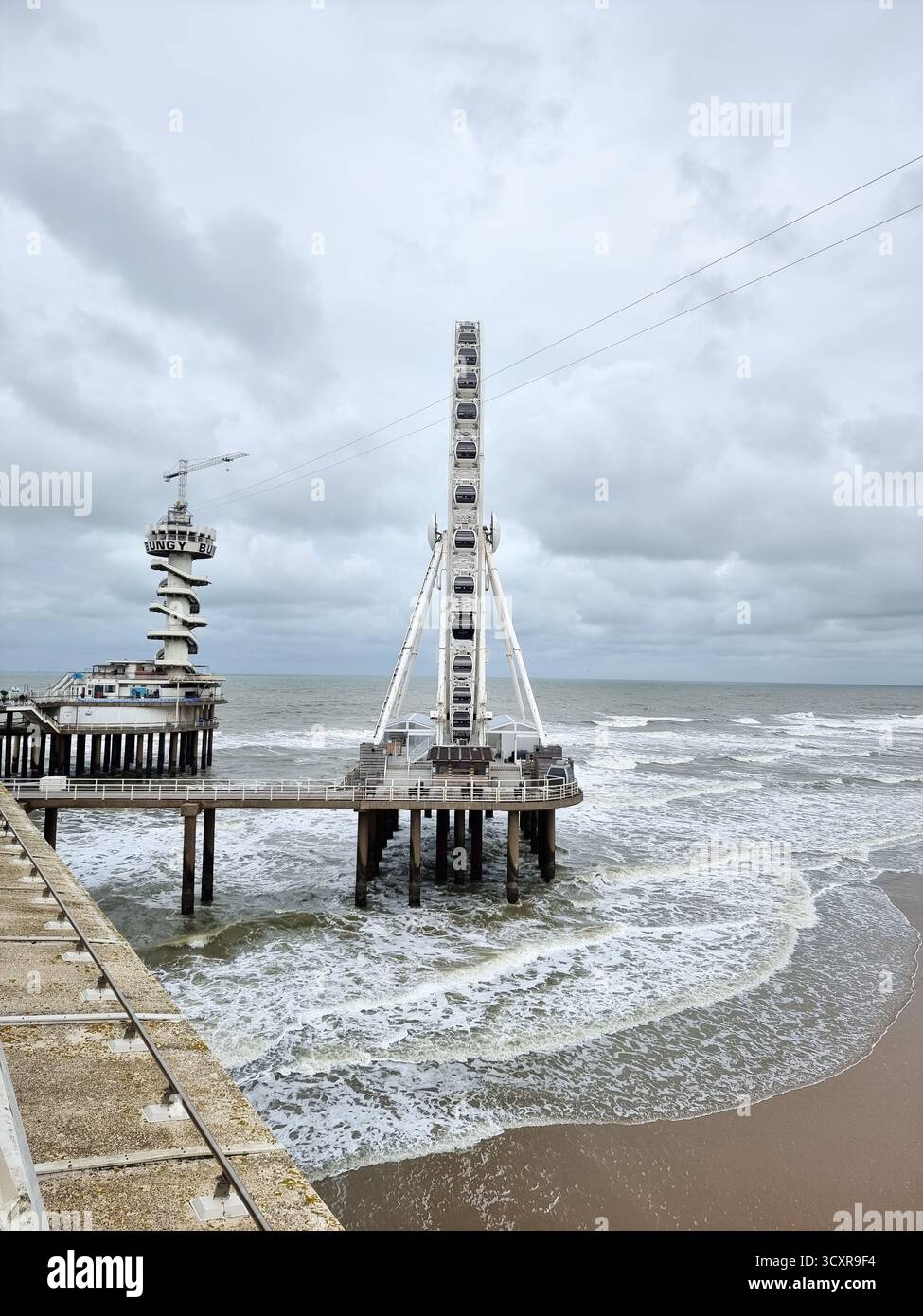 Side view of The Pier Skyview: ferris wheel in the North Sea on the pier in Scheveningen w/ clouds in autumn weather; Bungy Jump tower in background - Smartphone Captured Stock Image