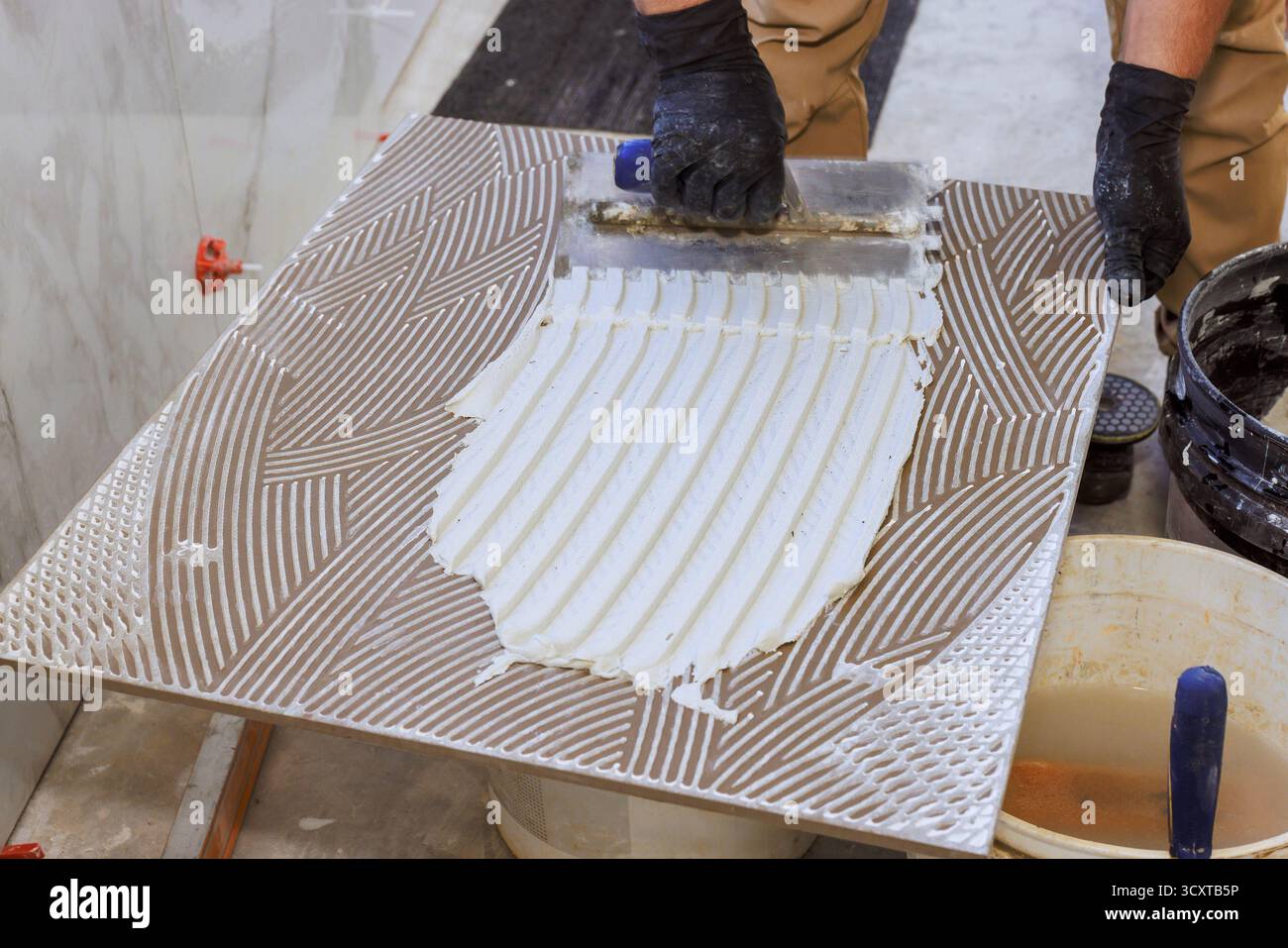 Worker uses trowel to spread adhesive on large tile ready for installation at building site. Stock Photo