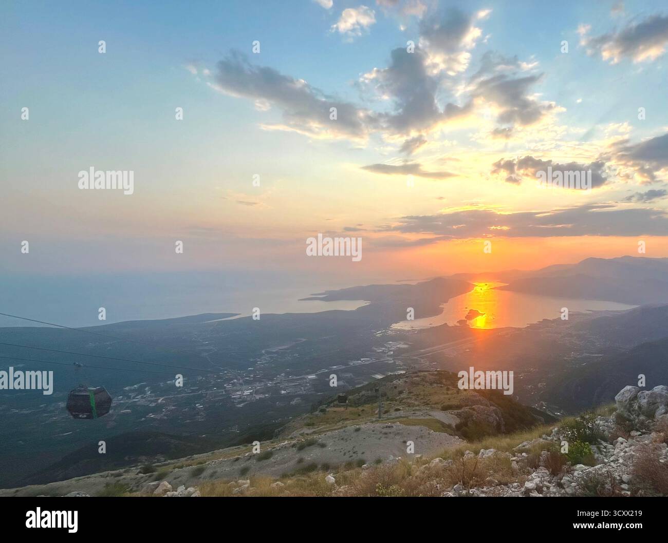 Sunset view over the coast from a mountain peak, with golden light reflecting on the sea and a cable car descending through the valley. - Smartphone Captured Stock Image