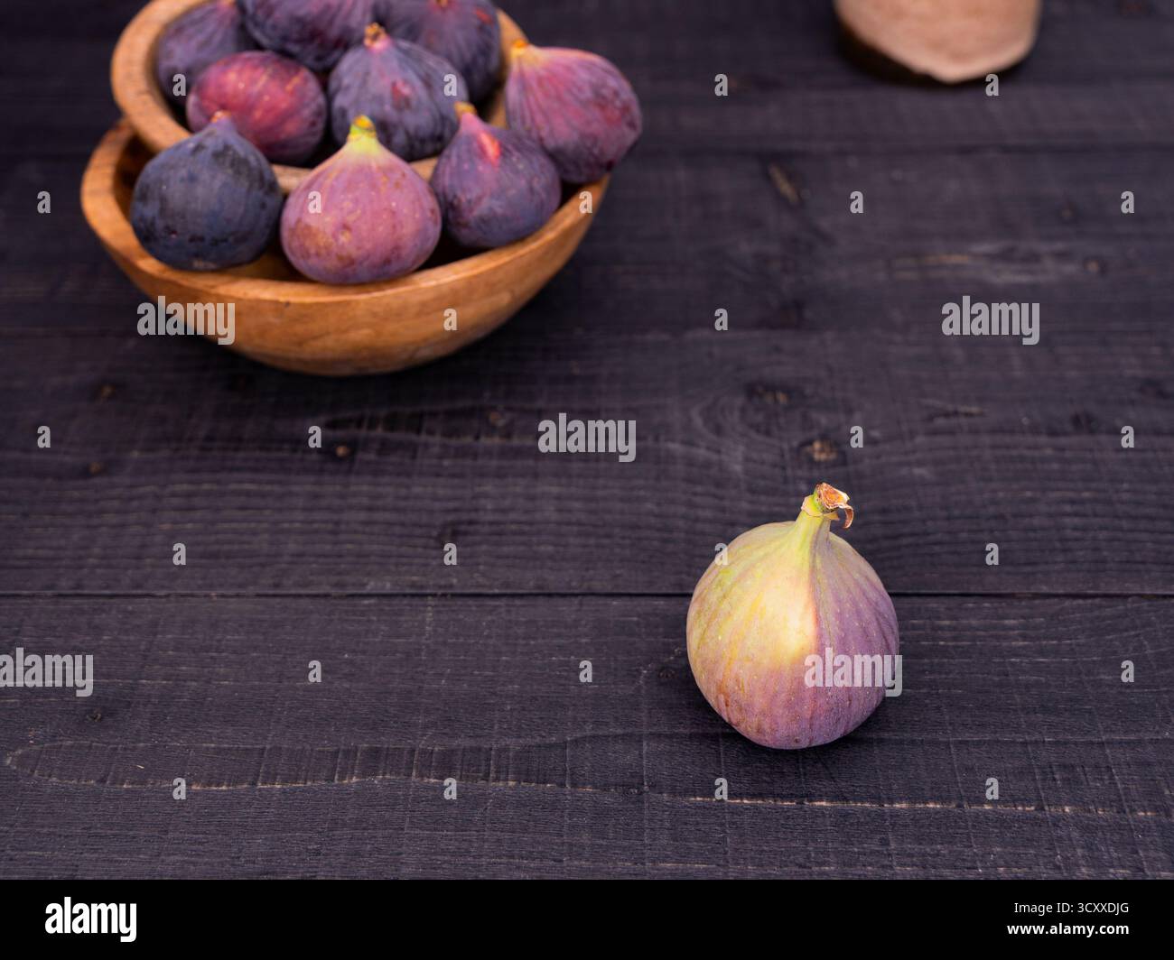 Fresh figs displayed on a black wooden table with one in front and more in wooden bowls, rustic and moody composition with vase in background Stock Photo