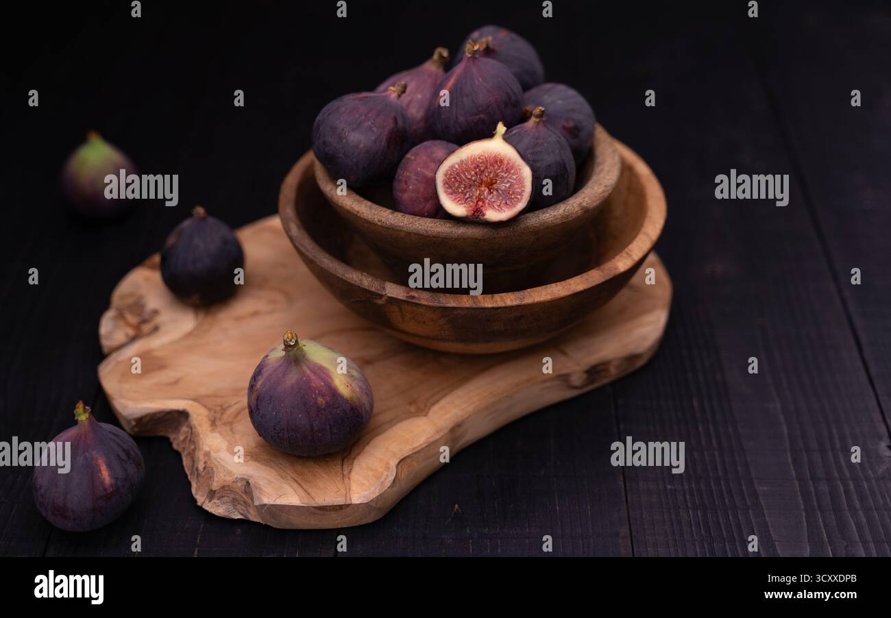 Half sliced fig in stacked bowls on wooden board with whole figs placed diagonally toward background, moody dark still life Stock Photo