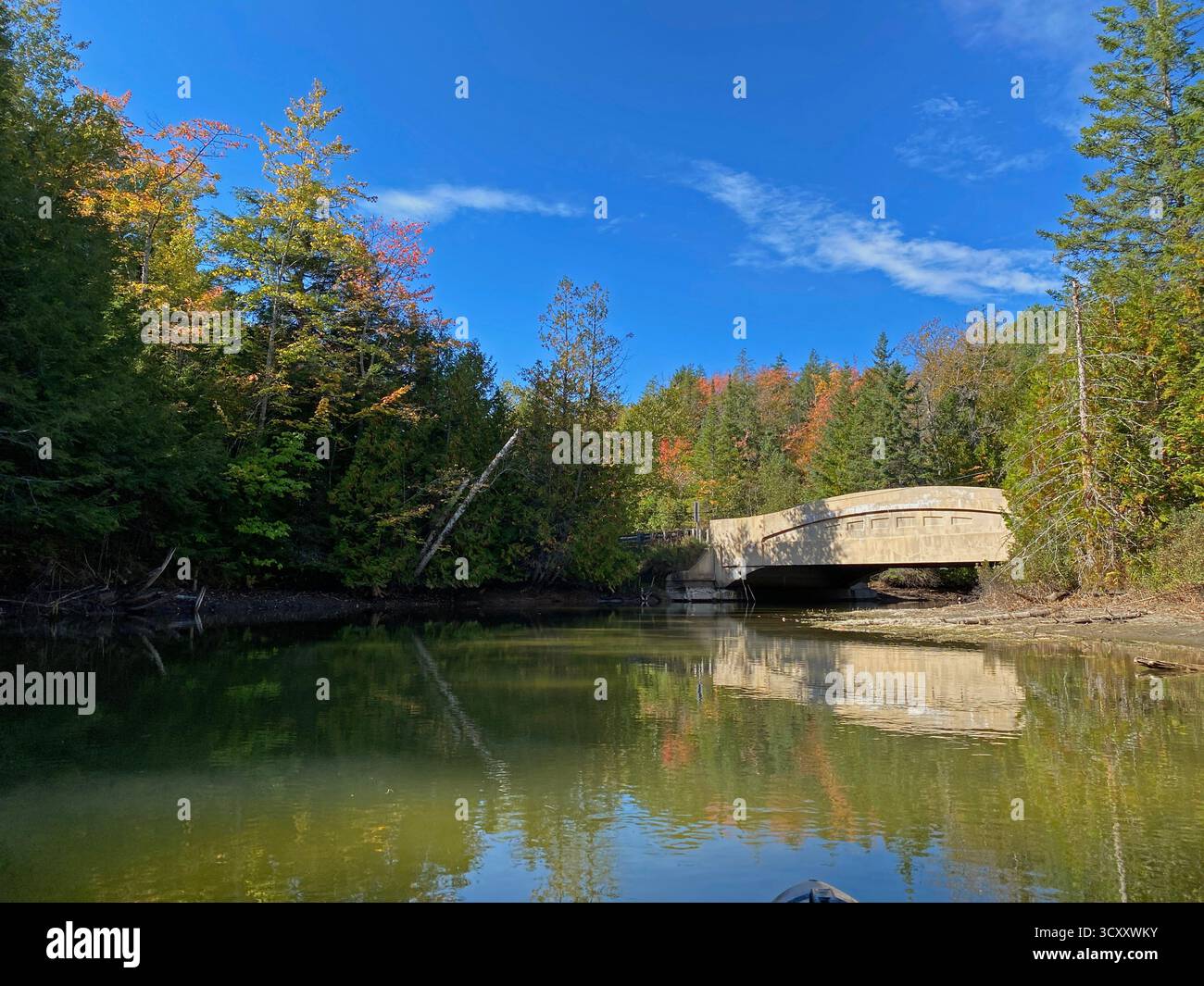 Kayaking down the Manistique River through a forest, lined with trees changing colors in the fall, with a low bridge ahead, in Germfask, Michigan - Smartphone Captured Stock Image