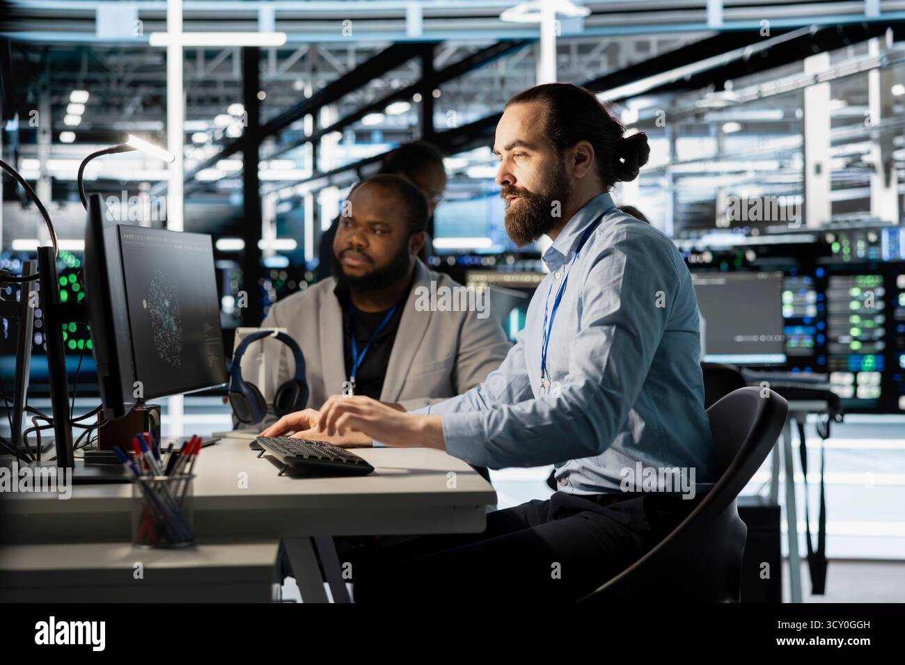 Team of programmers using computer in server farm, analyzing data. Men working in data center office examining infrastructure using device, ensuring system integrity and security Stock Photo